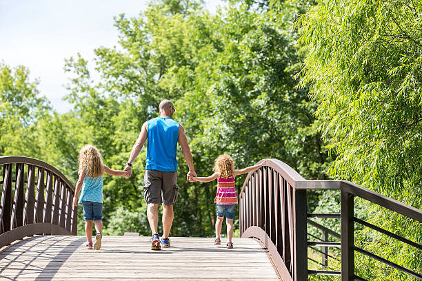 istock Father & Daughters Walking Across Bridge At Park 589427308