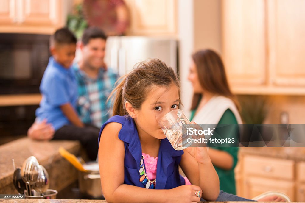 Hispanic girl and family cooking dinner together in home kitchen. Loving Hispanic family cooks dinner together in home kitchen.  Daughter foreground drinks water in clear glass. Drinking Water Stock Photo