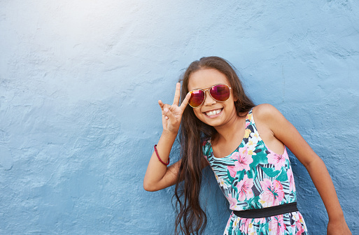 Portrait of cute little girl with sunglasses gesturing peace sign. Preteen girl standing against blue wall with copy space.