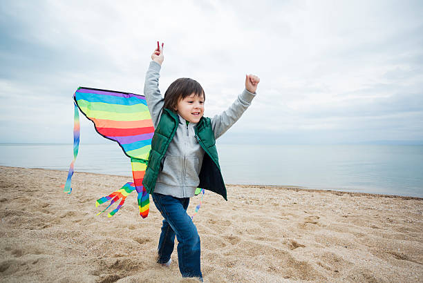 Joyful young boy running along the beach and flying a colorful kite on a bright windy day. Fun outdoor activity for kids by the sea