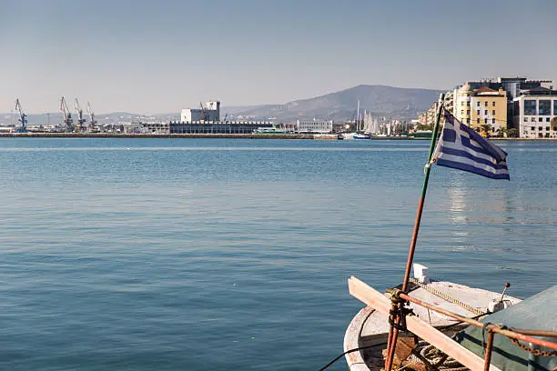 In Greece, on the Aegean sea's Pagasetic Gulf, a boat bearing the Greek flag, is docked at Volos quay. Mount Pelion, some buildings and the port  are visible in the background.