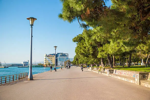Beautiful promenade in Volos, Greece, by the Aegean sea's Pagasetic Gulf. The Pelion mountain range and the port are visible in the far background and so are some unrecognizable people walking.