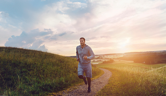 Male runner makes his way forward on small road through a meadow. Sun sets in the horizon. The runner wears sports clothing and running shoes.