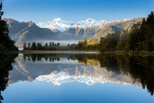 \nReflections in the still waters of Lake Matheson on the South Island of New Zealand. Early morning mist remains as the rising sun illuminates the rain forest surrounding the lake.\n\nLake Matheson is a popular destination for tourists visiting the West Coast of New Zealand. It is well known for its reflections of the surrounding forest and mountains, including New Zealand's tallest mountain Mount Cook.