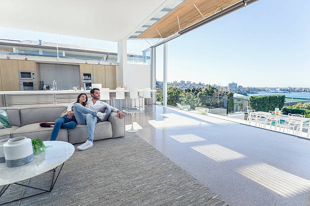 Couple sitting in a modern open plan house. Couple sitting in a modern open plan house. They are sitting on a sofa with a kitchen behind them. They are looking at the beautiful water view from their house. They are attractive, smiling and happy. luxury lifestyle city stock pictures, royalty-free photos & images