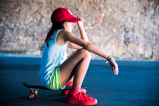 Side view of a cute little girl with red cap and red sneakers sitting on her black skateboard with red wheels. One arm is stretched forward and with one arm she is holding her cap and she is looking ahead.