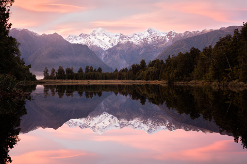 A spectacular pink sunrise reflects in the still waters of Lake Matheson together with snow capped mountains and the rain forest surrounding the lake. 