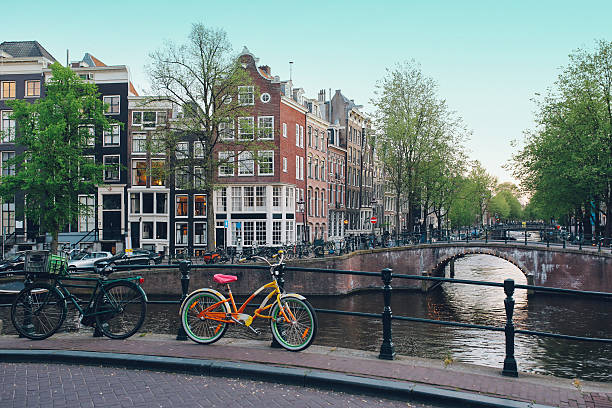 Nice buildings in the Amsterdam city center on a bright summertime day with bicycles parked by the canal and people walking by as the sun sets behind the scene.