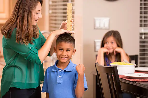 Latin descent mother measures her son's growth with a yard stick at home setting. Daughter is in background. Single mother.