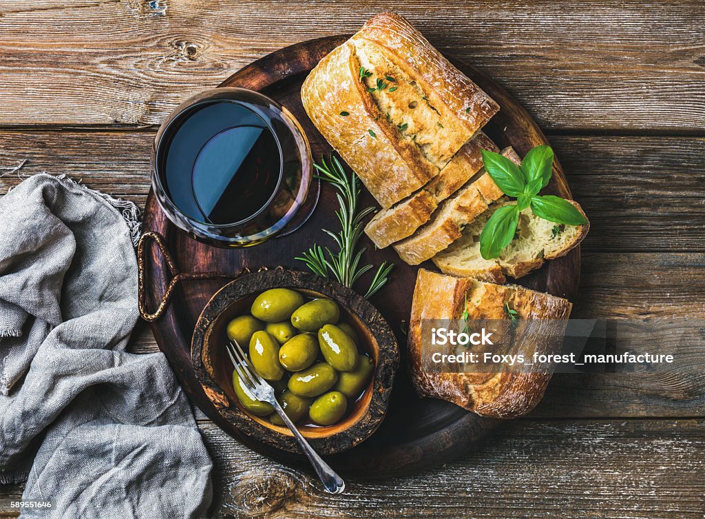 Red wine glass, green olives and ciabatta over wooden background Wine snack set. Glass of red wine, green mediterranean olives, freshly baked ciabatta bread in dark wooden plate over rustic wooden background. Top view, horizontal composition Olive - Fruit Stock Photo