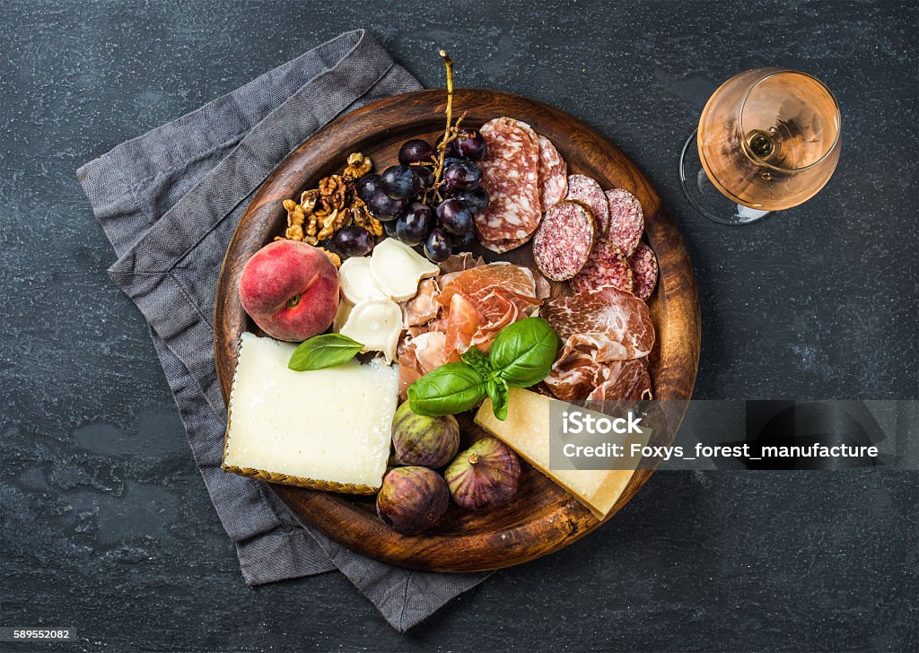 Italian antipasti snack for wine on wooden tray, dark background Italian antipasti snack for wine. Prosciutto di Parma, salami, cheese variety, figs, grapes, peach, walnuts and fresh basil on wooden serving tray with glass of Rose, dark grunge background. Top view Alcohol - Drink Stock Photo