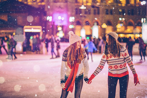 With bestie for Christmas Friends having so much fun while ice skating at night. Wearing warm clothing. City is decorated with christmas lights. ice skating photos stock pictures, royalty-free photos & images