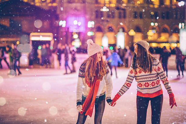 With bestie for Christmas Friends having so much fun while ice skating at night. Wearing warm clothing. City is decorated with christmas lights. skate rink stock pictures, royalty-free photos & images