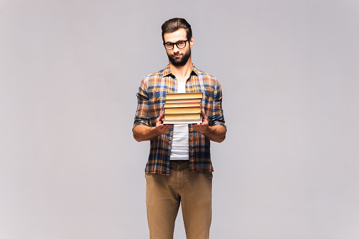 Studio shot of frustrated young man in eyeglasses and casual wear carrying book stack and looking frustrated