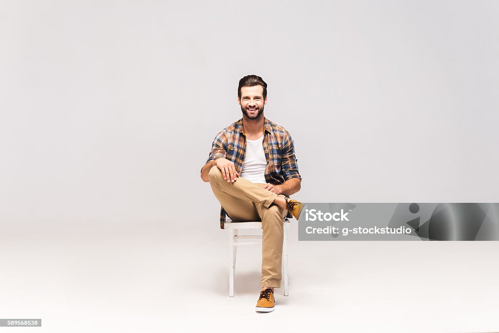 Casually handsome. Studio shot of handsome young man sitting on the chair and looking at camera with smile Sitting Stock Photo