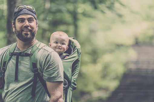 DSLR Picture of a father Backpacking or Hiking with a Baby boy inside a baby carrier in Forest on a nice summer day.