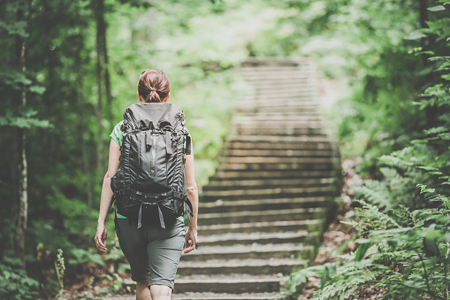 DSLR picture of a woman hiking or trekking with backpack on a footpath in forest on a nice day of summer. There is wood and rock stairs to climb in front of her.