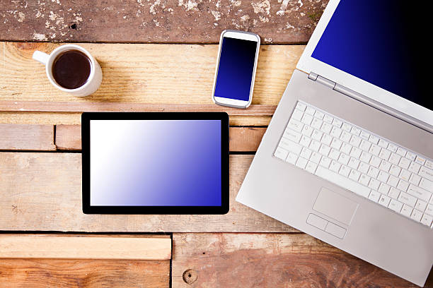 Overhead view of business person's 'tools.'  Laptop, cell phone, digital tablet, and coffee cup. Blank, blue background on screens. Unique wooden table background.  Copyspace.