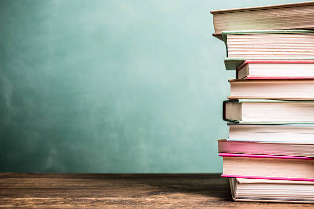 Textbooks stacked on school desk with chalkboard background. It's back to school time!  A large stack of textbooks to side makes frame composition.  The pile of objects lies on top of a wooden school desk with a green chalkboard in the background.  The blank blackboard in the background makes perfect copyspace!  Education background themes. textbook stock pictures, royalty-free photos & images