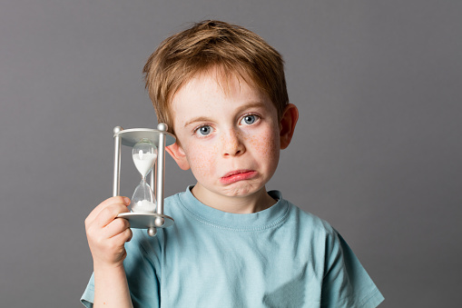 worried young child with sad blue eyes and a pouting mouth holding an egg timer, showing his disappointment in growing up for time concept, grey background