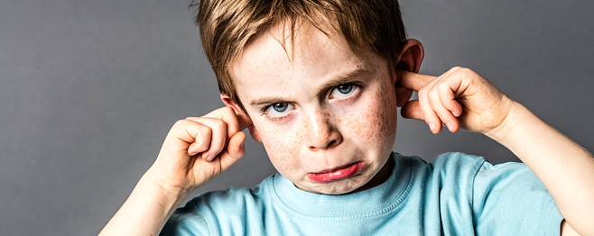 closeup of displeased little boy with red hair and freckles ignoring parents scolding, blocking his ears with fingers against education problems, grey background