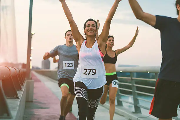 Runners running marathon in the city. They are running with arms raised over the bridge at sunset. Selective focus on beautiful brunette woman running and smiling. Wearing numbers on their sport clothes.  Running through the finish with arms raised.