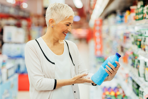 Mature smiling woman shopping in local supermarket. She is shopping groceries. Standing by produce stand and reading nutrition label on dairy product.