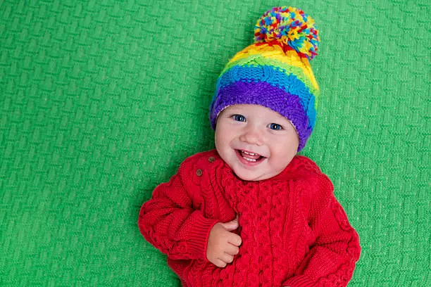 Cute baby in warm wool knitted hat on a red blanket. Autumn and winter clothing for young kids. Colorful knitwear for children. Adorable little boy ready for a walk on a cold fall day.