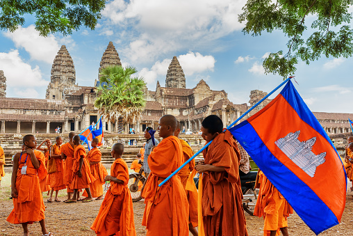 Row of Thai monks walking along people is   getting food donations  at  ceremony of Makha Bucha Day in Sangkhla Buri in northwest of Kanchanaburi province of Thailand in early morning close to sunset. People are wearing mostly traditional clothing and are standing in main street leading down to Mon Bridge and lake. People are waiting to give food donations to monks when they pass along street down to bridge.