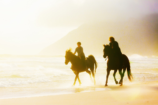Two women horseback riders on a  beach on a windy winter's afternoon, are almost silhouetted against the choppy sea and misty, gold-tinged sky. Ample copy space over the sea and misty mountain.