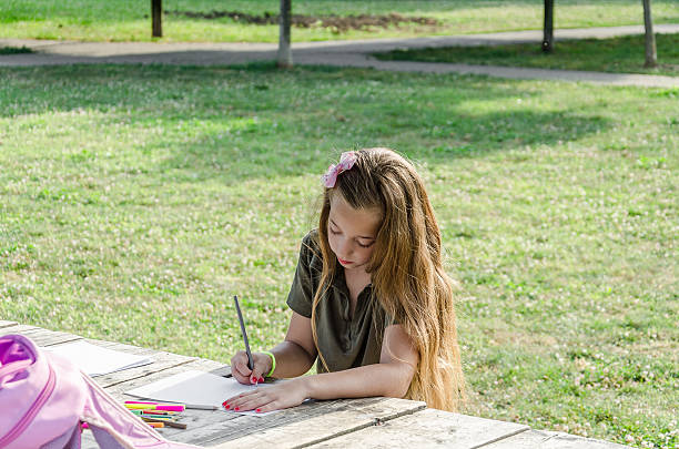 小さな女の子は木のテーブルの上に公園で描きます - schoolgirl little girls crayon human face ストックフォトと画像