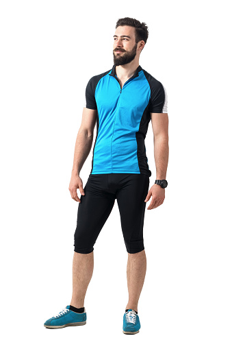 Young bearded cyclist guy in blue jersey t-shirt and leggings relaxed smiling and looking away. Full body length portrait isolated over white studio background.