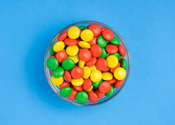 A bowl of yellow, green, and orange, hard chocolate covered candies. Shot from above on a blue background.