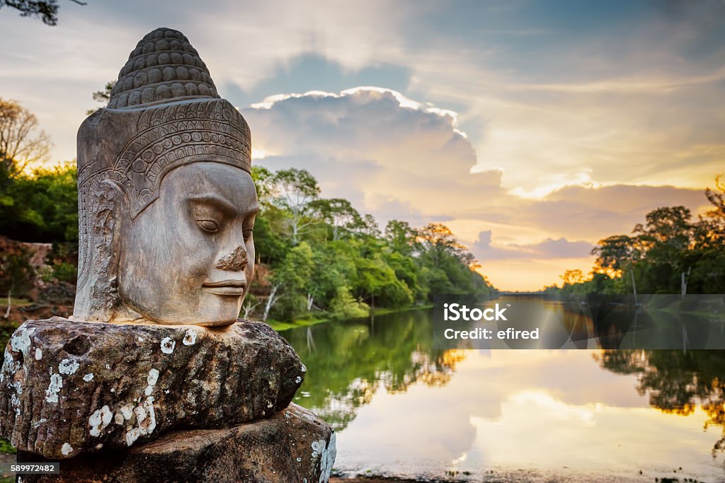 Stone face Asura and sunset over moat. Angkor Thom, Cambodia Stone face Asura on causeway near South Gate of Angkor Thom in Siem Reap, Cambodia. Beautiful sunset over ancient moat in background. Mysterious Angkor Thom is a popular tourist attraction. Cambodia Stock Photo