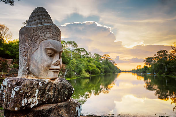 Stone face Asura and sunset over moat. Angkor Thom, Cambodia Stone face Asura on causeway near South Gate of Angkor Thom in Siem Reap, Cambodia. Beautiful sunset over ancient moat in background. Mysterious Angkor Thom is a popular tourist attraction. cambodia stock pictures, royalty-free photos & images