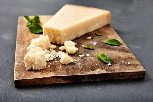 Pieces of parmigiano reggiano or parmesan cheese on wood board on stone background. Parmesan is hard cheese uses in pasta dishes, soups, risottos and grated over salads.