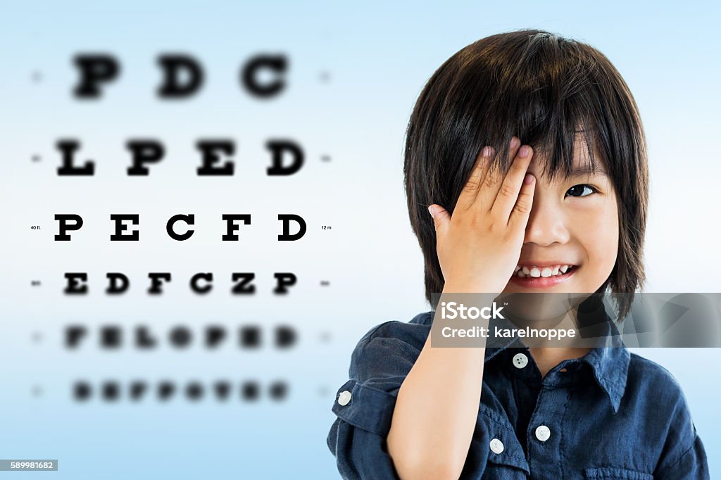 Cute asian boy doing eye test. Close up portrait of cute little asian boy doing eye test.Kid closing one eye with hand against alphabetical out of focus test chart in background. Eye Exam Stock Photo