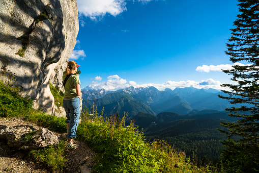 Woman standing and looking at the beautiful Alpine Logar Valley mountains from entrance of Potocna Zijavka Cave in Slovenia, close to the border with Austria.  Potocna Zijavka Cave at Olseva mountain is archaeological site from stone age and is on pleasant hiking bear trail.