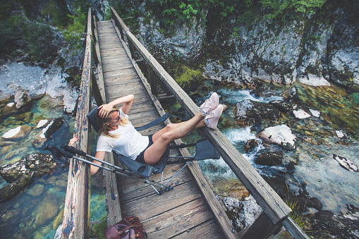 Girl sitting on the camp chair on the bridge over the mountain stream in the forest