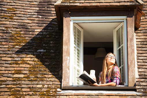 Young woman standing on rooftop window. Holding a book and a coffee cup. Enjoying sunny  morning, eyes closed. With long hair, eyeglasses and checkered shirt. Old architecture building rooftop around.