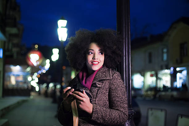 Portrait of smiling young woman using smart phone on street by night. Woman with curly hair, wears purple pullover and gray coat. Building exterior and defocused street lights as background.