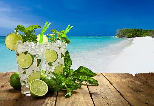 Two mojito cubano drinks on rustic wood table against tropical beach
