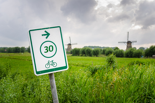 White sign, reading the route number and the direction, on a bicycle path in the countryside of the Netherlands, Some windmills are visible in background.