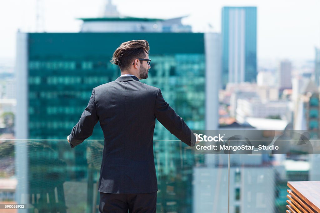 Young Businessman Looking at City Portrait of young businessman standing on balcony and looking at city view Men Stock Photo