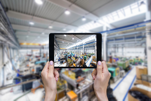 Horizontal color image of cropped hands of unrecognizable person taking pictures of factory with machines on a digital tablet. Female hands holding a digital tablet in a modern plastic production line. Ordering on-line from injection moulding factory on a touchscreen tablet computer. Large factory, industrial machines, robots and manufacturing equipment arranged in background.