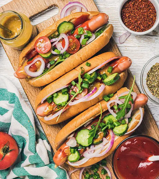 Homemade hot-dogs on wooden serving board with fresh vegetables, spices, ketchup and mustard over white painted background, top view