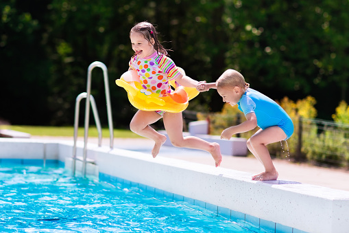 Happy little girl and boy holding hands jumping into outdoor swimming pool in a tropical resort during family summer vacation. Kids learning to swim. Water fun for children.