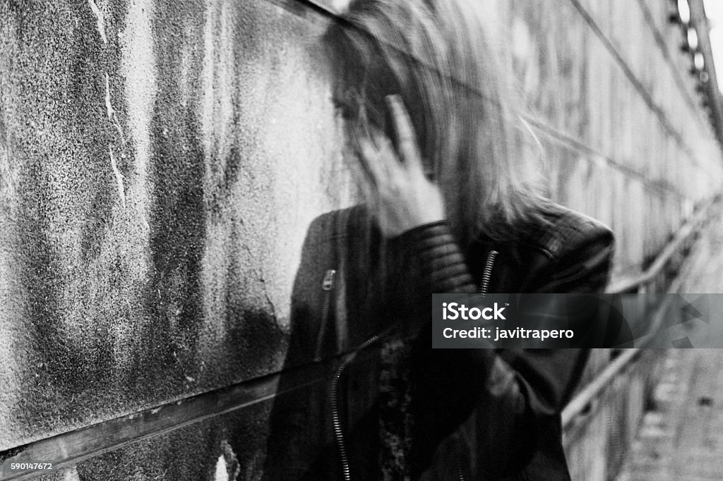 Young woman suffering from disorientation, confusion, or sadness. Young woman suffering from a severe disorientation, confusion, or sadness outdoors, in front of a wall. Converted to black and white, grain added, blurry, slightly out of focus. Addiction Stock Photo