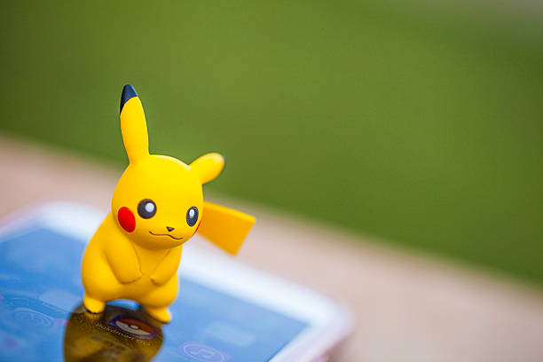 Peyton, Colorado, USA - August 17, 2016: A horizontal shot of the Pokemon Go character Pikachu, standing on top of an Apple iPhone 6 Plus. The Pokemon Go icon can be seen on the phone screen. The character and phone are on a wall outdoors, in front of a defocused grassy area in Peyton, Colorado. The figurine is made by Tomy. 