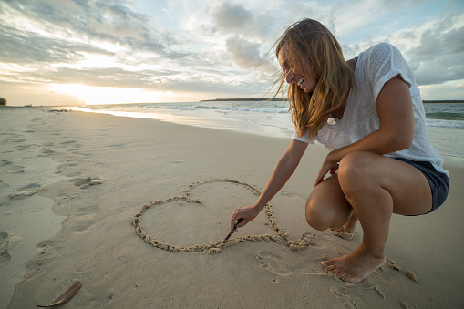 Young woman on the beach drawing a heart shape on the sand at sunset.